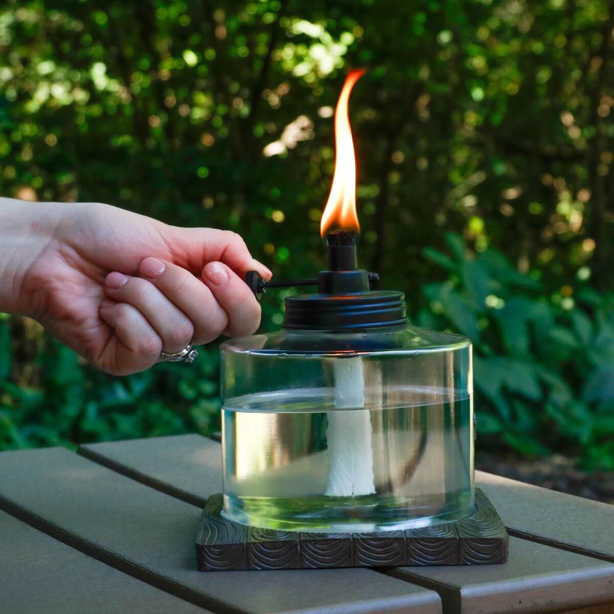 A hand lights a clear TIKI® tabletop torch with a visible flame, set on an outdoor table with greenery in the background.