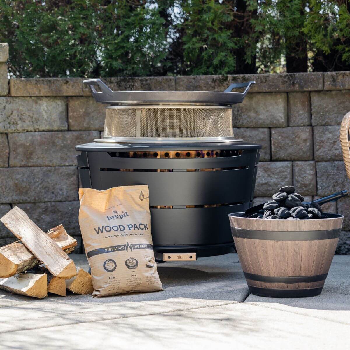 TIKI® Fire Pit on patio with wood pack, firewood, and decorative barrel of stones; outdoor setting with stone wall.