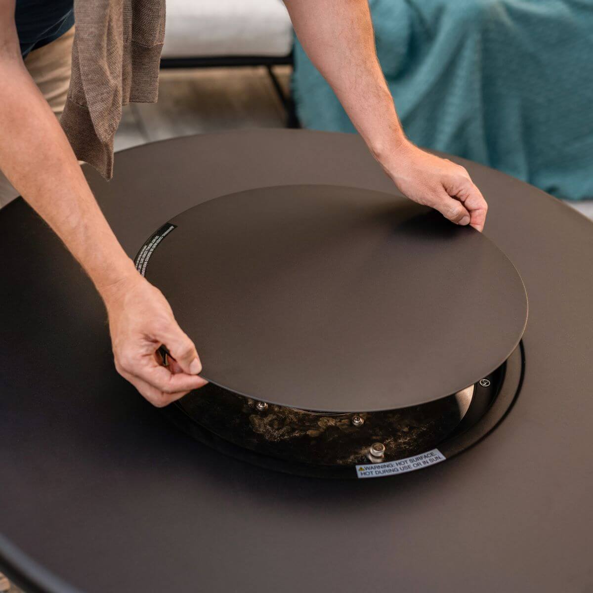Person lifting the lid off a TIKI® Brand fire pit, revealing the burner inside.