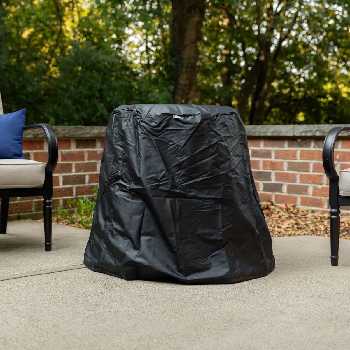 Black TIKI fire pit cover protects your outdoor fire pit from weather; shown on a patio between two chairs.