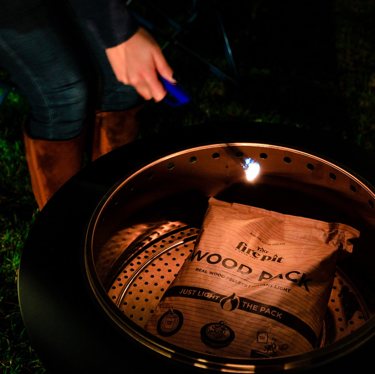 Person lighting a TIKI® Wood Pack in a fire pit outdoors; easy-start wood pellets for quick, clean fire.