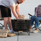 Round black metal fire pit with slatted sides, set on a patio. Person adds wood pack fuel. Two people seated nearby.