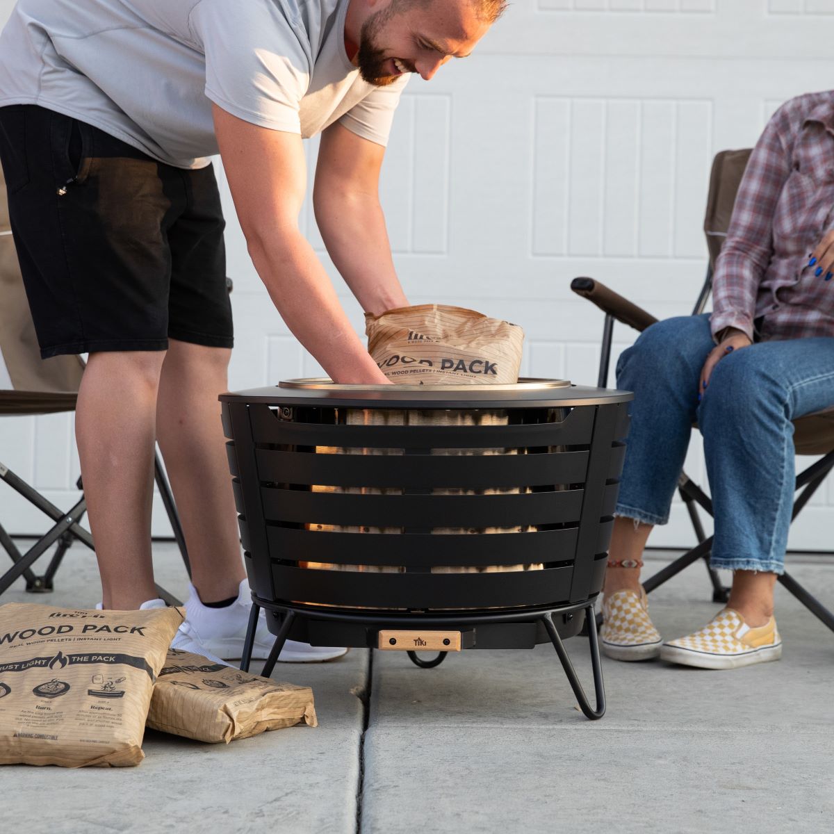 Round black metal fire pit with slatted sides, set on a patio. Person adds wood pack fuel. Two people seated nearby.