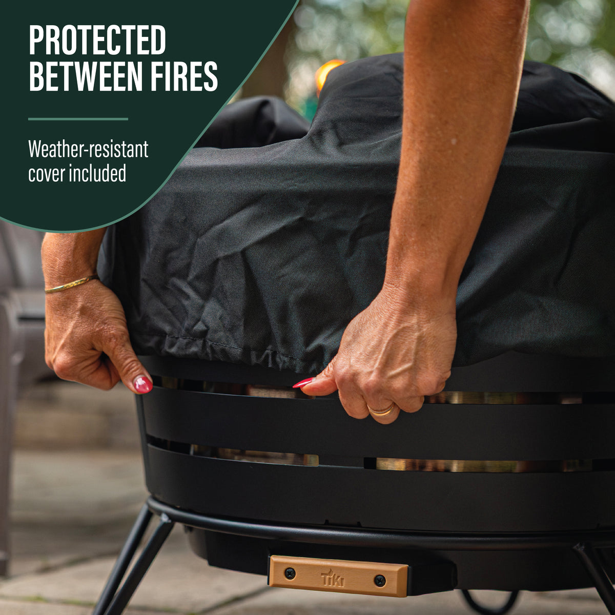 Round black fire pit with a weather-resistant cover being placed on top; Tiki brand label visible.