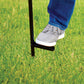 A person uses their foot to press a TIKI torch stake into grass for secure outdoor placement.