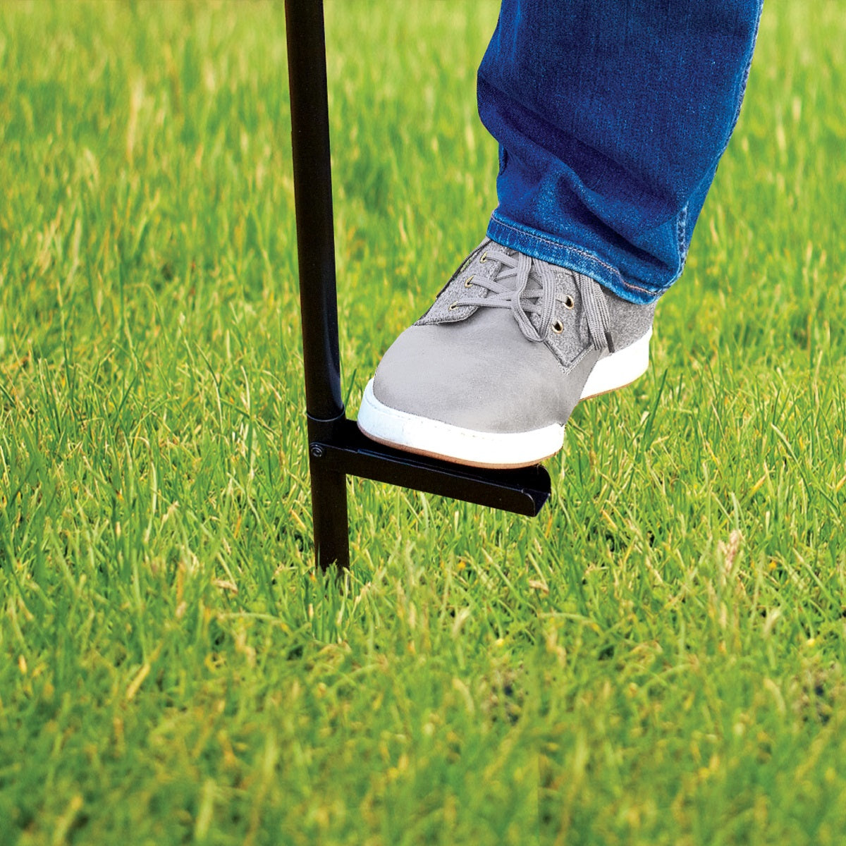 A person uses their foot to press a TIKI torch stake into grass for secure outdoor placement.
