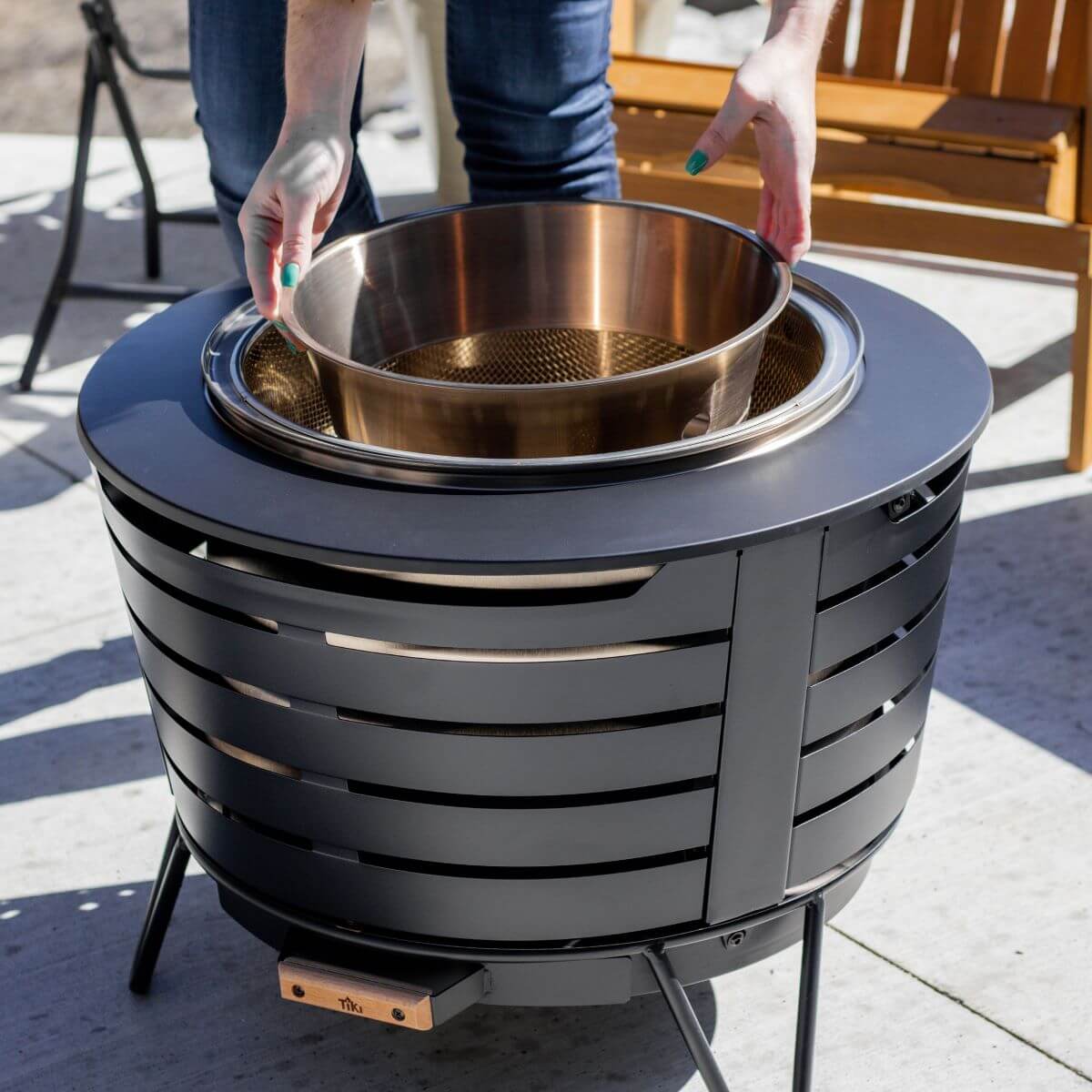 Person placing a metal fire bowl into a TIKI patio fire pit on a concrete outdoor surface.