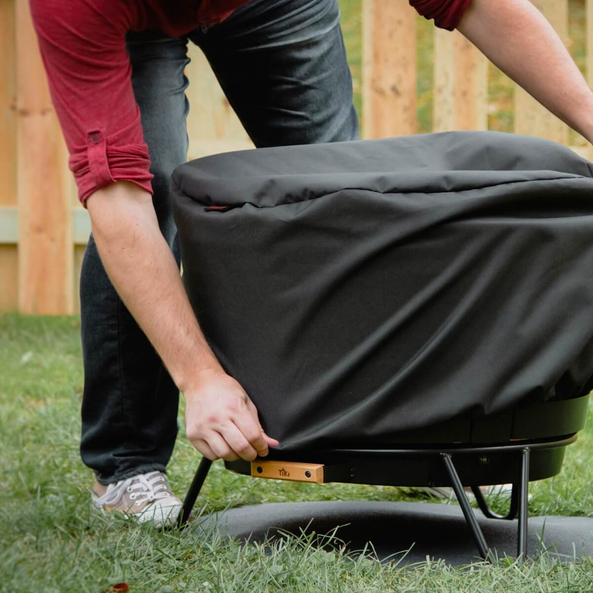 TIKI Brand Reunion Smokeless Fire Pit Replacement Cover and Hardware, hands pulling the cover down over a fire pit's base
