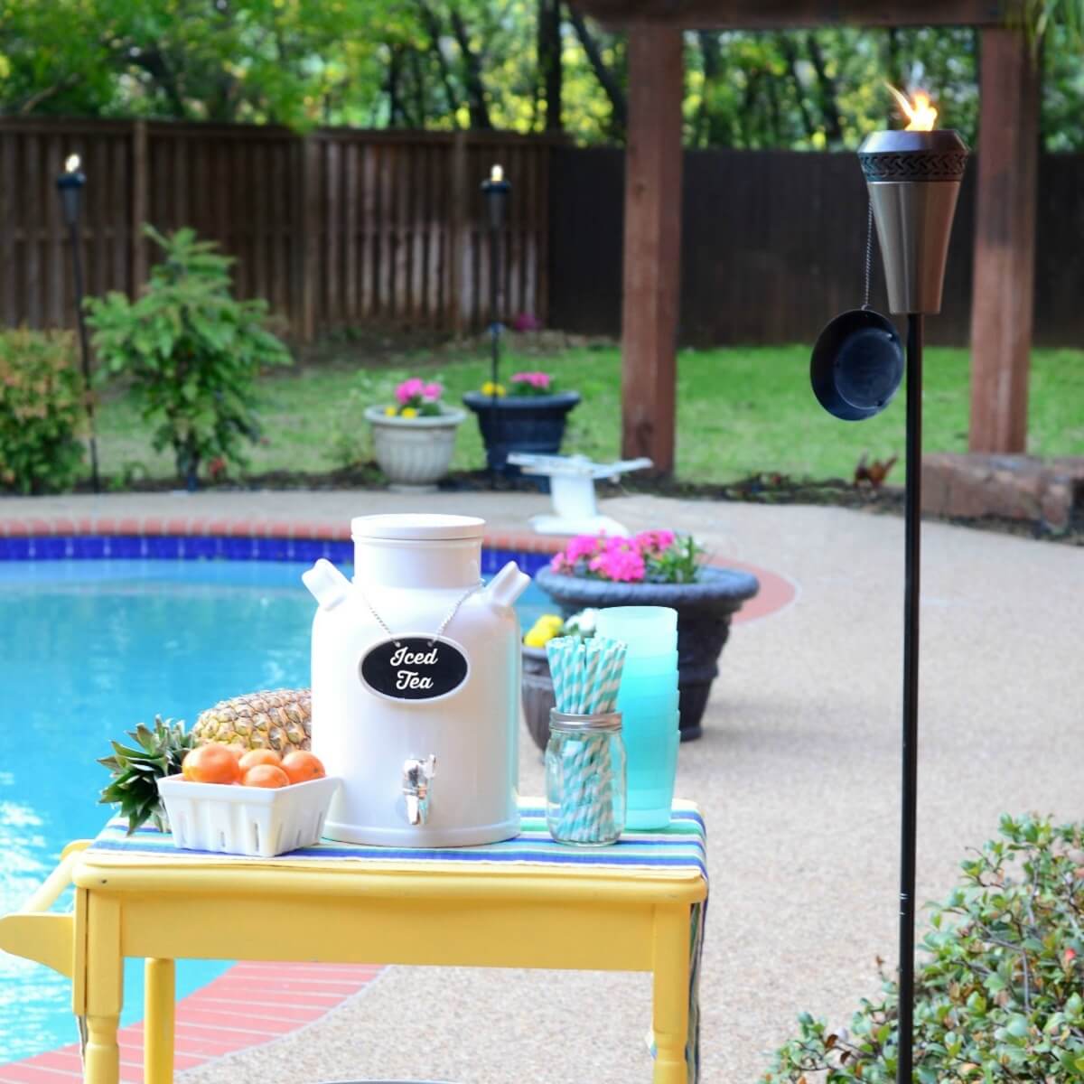 Outdoor poolside setup with TIKI torch, iced tea dispenser, fruit, cups, and straws on a yellow table.