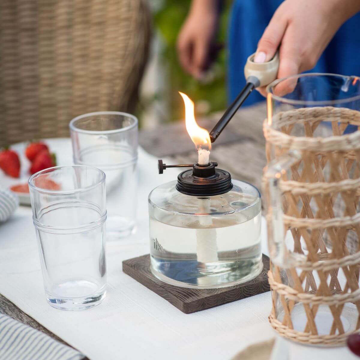 A hand lights a TIKI® table torch on an outdoor dining table set with glasses and a woven lantern.
