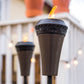 Close-up of a lit TIKI® torch on a patio, with a second torch and string lights in the background.