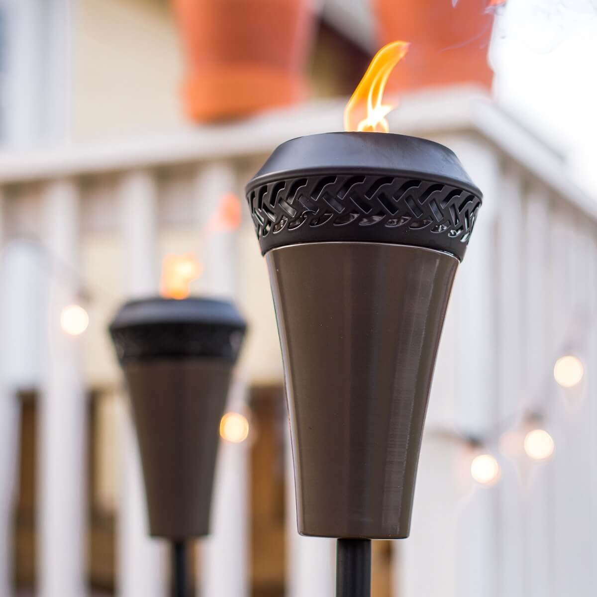 Close-up of a lit TIKI® torch on a patio, with a second torch and string lights in the background.