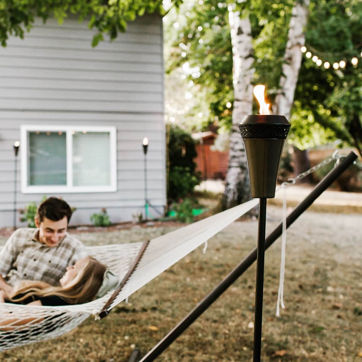 A TIKI® torch glows beside a hammock in a cozy backyard, creating a relaxing outdoor ambiance near a home.