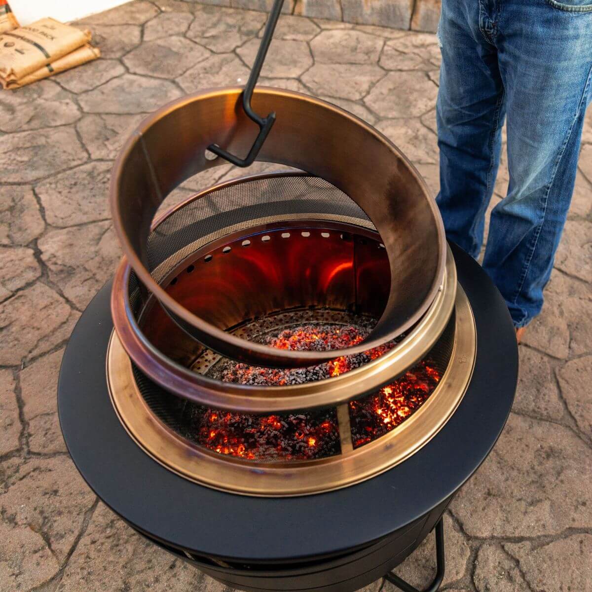 Bronze fire pit with glowing embers, black base, and mesh screen.
