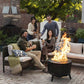 Four people enjoy drinks around a TIKI® fire pit on a cozy patio with outdoor seating and lush greenery.