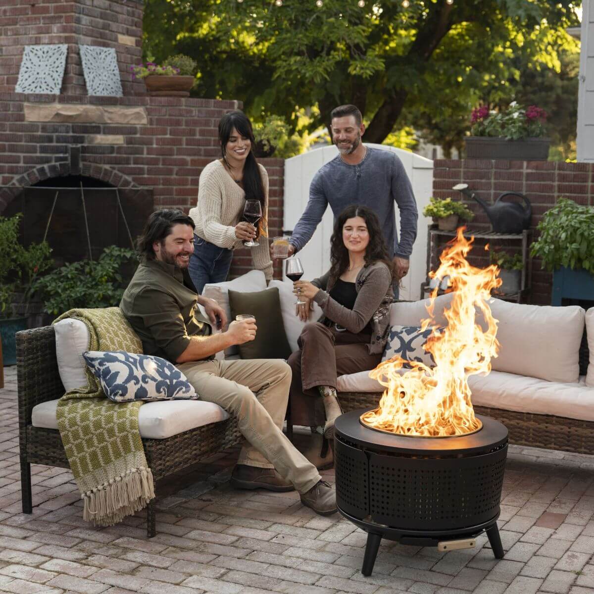 Four people enjoy drinks around a TIKI® fire pit on a cozy patio with outdoor seating and lush greenery.