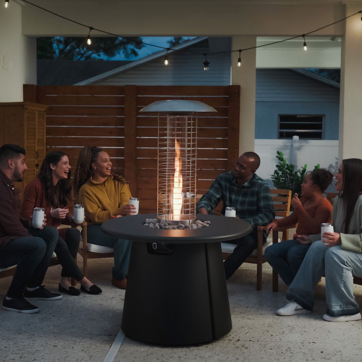 A group enjoys drinks around a TIKI® Brand fire pit on a patio with string lights, creating a cozy outdoor gathering.