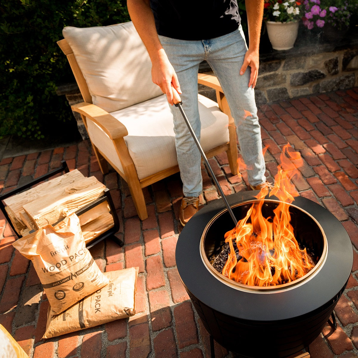 Person tending a TIKI fire pit on a brick patio, with wood packs and logs nearby; outdoor seating and flowers in background.