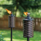 Two woven-style TIKI® torches with flames lit, set outdoors in a green backyard with a wooden fence.