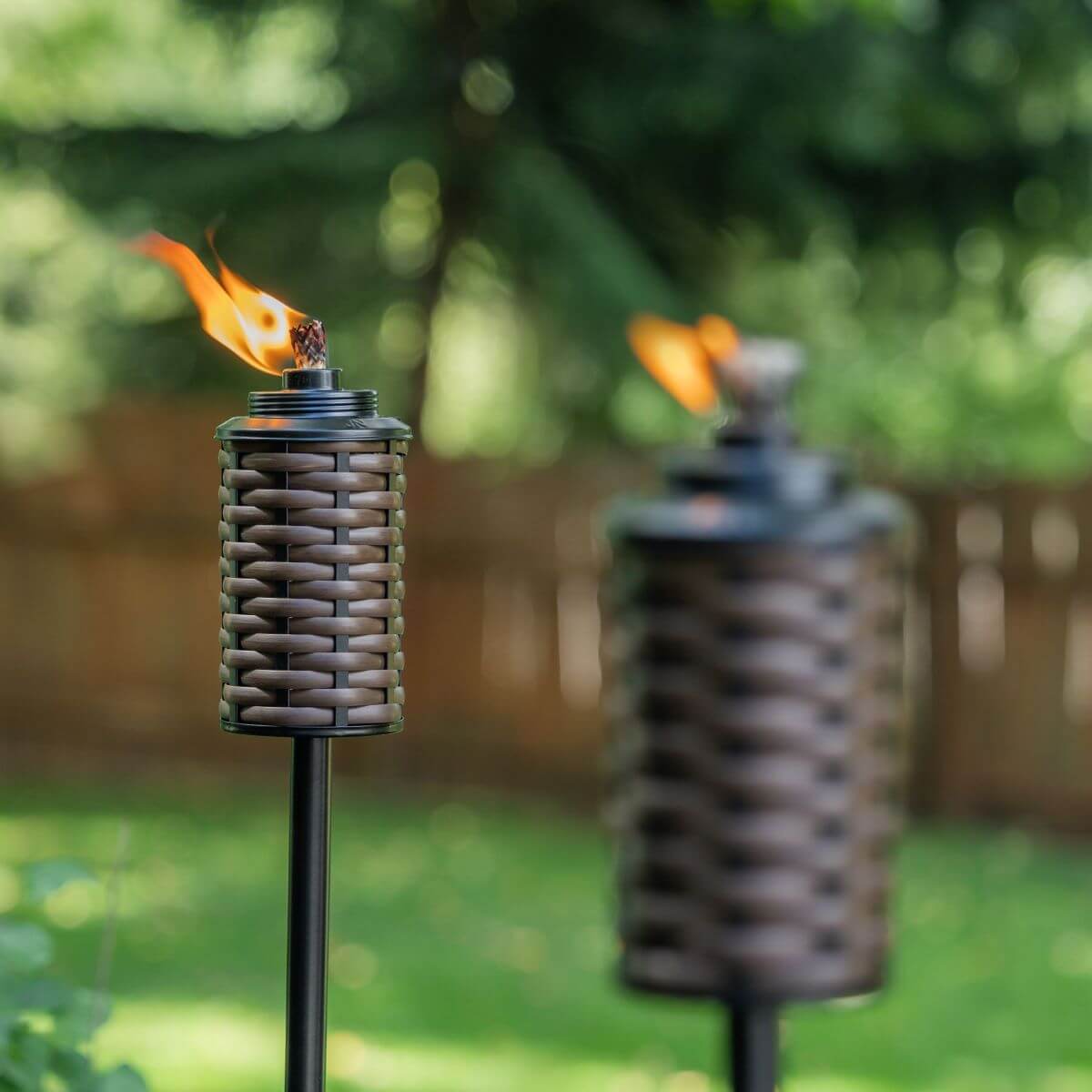 Two woven-style TIKI® torches with flames lit, set outdoors in a green backyard with a wooden fence.
