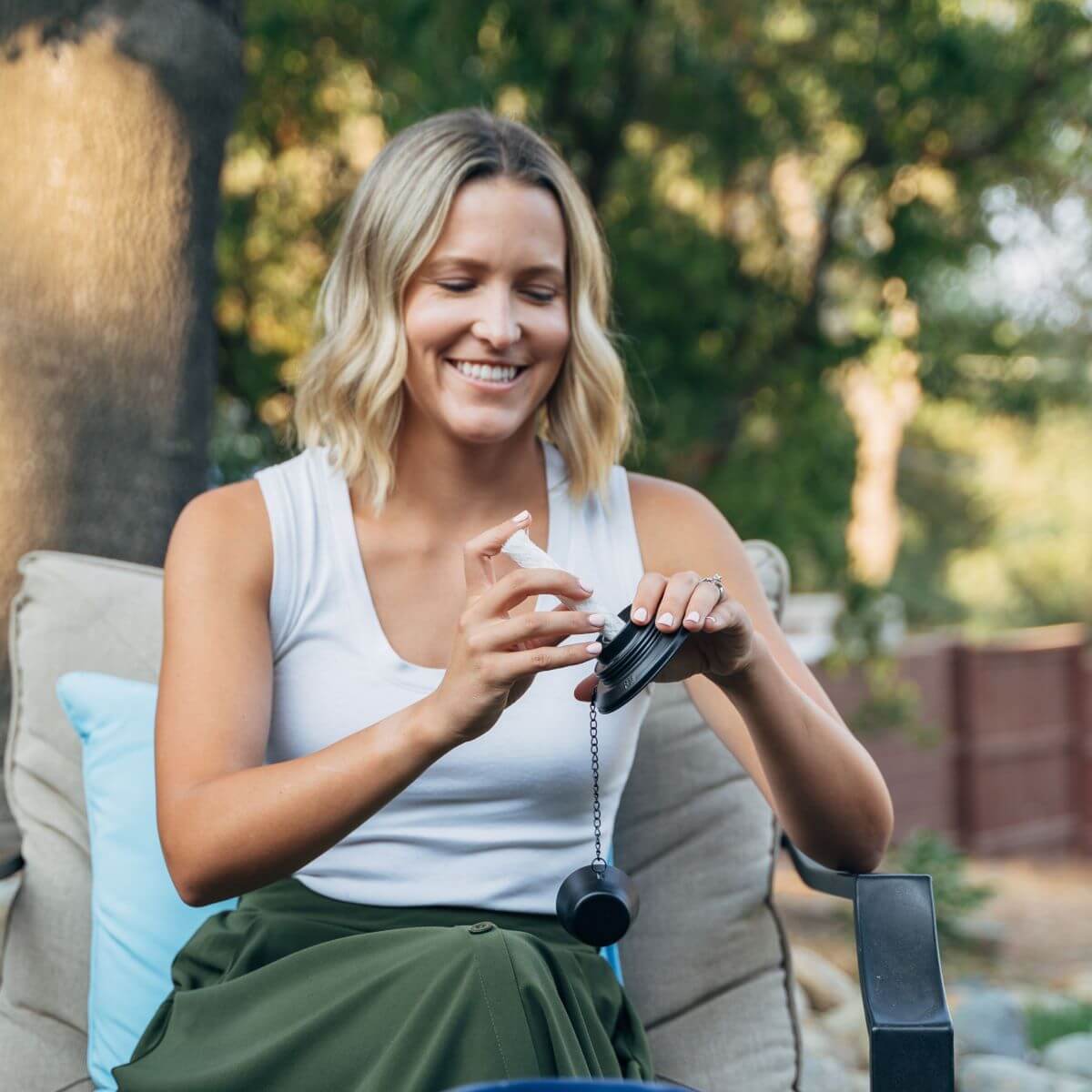 Woman sitting outdoors, applying TIKI Brand BiteFighter repellent to a patio accessory for mosquito protection.
