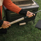 Person lighting a TIKI Brand fire pit with a long lighter outdoors on grass, showing safe fire starting technique.