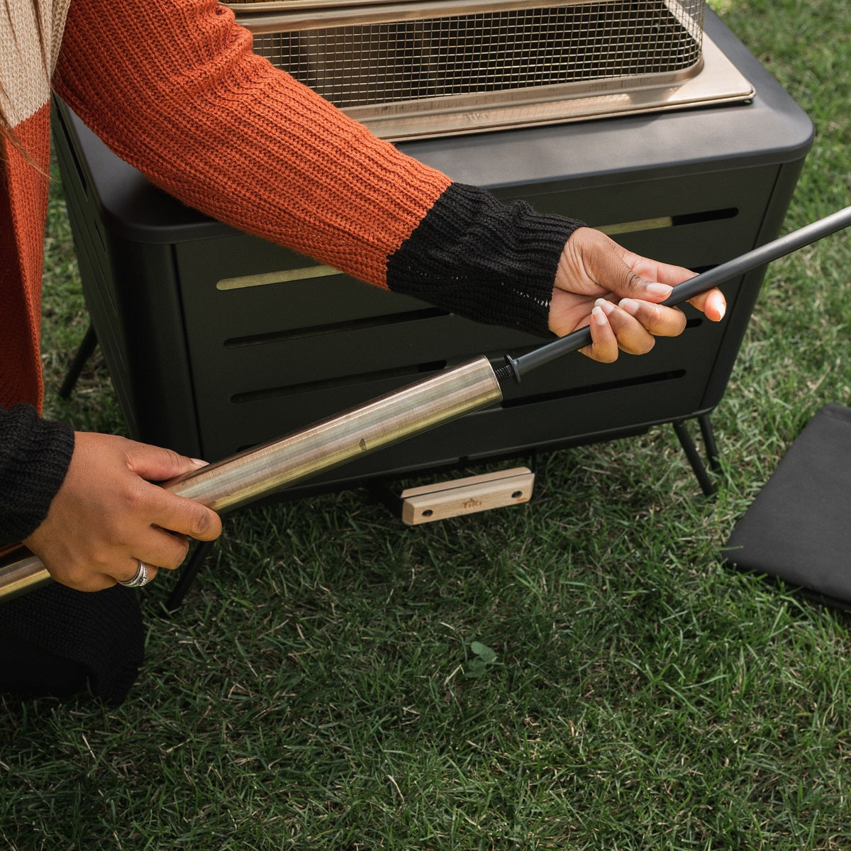 Person lighting a TIKI Brand fire pit with a long lighter outdoors on grass, showing safe fire starting technique.