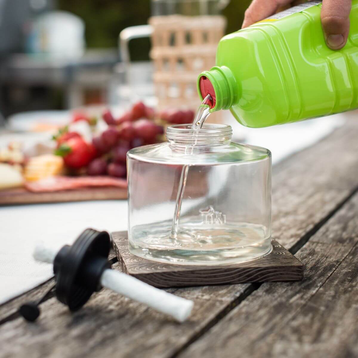 A hand pours TIKI torch fuel into a clear torch canister on a wooden table; wick and cap are beside it.