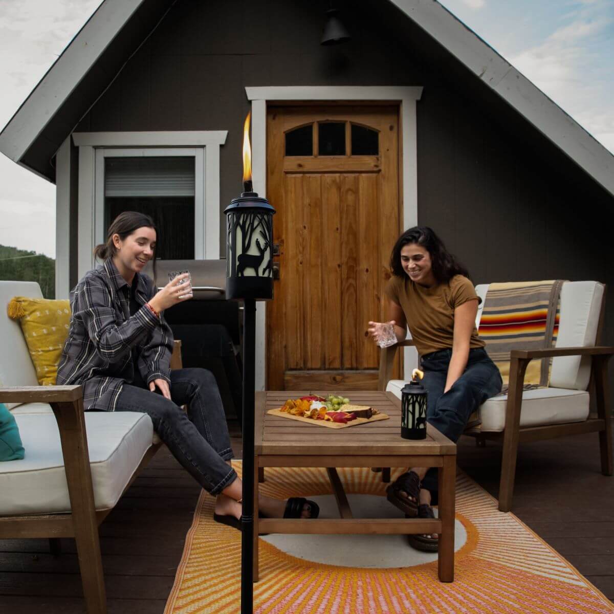 Two people relax on a patio with TIKI Brand torches and tabletop torch, enjoying drinks and snacks on outdoor furniture.