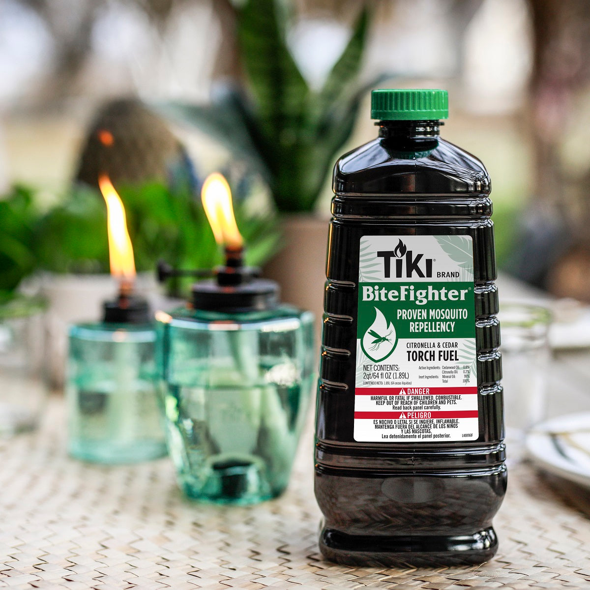 TIKI BiteFighter Torch Fuel bottle on a patio table with two lit torches in the background, outdoor setting.