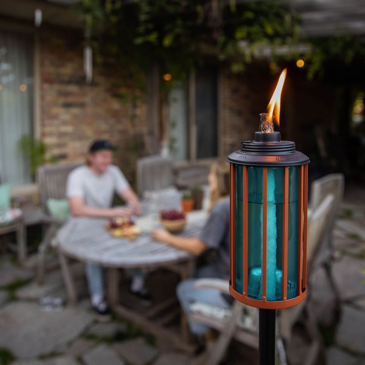 A TIKI® Brand torch burning in a backyard, with people enjoying snacks at a patio table in the background.