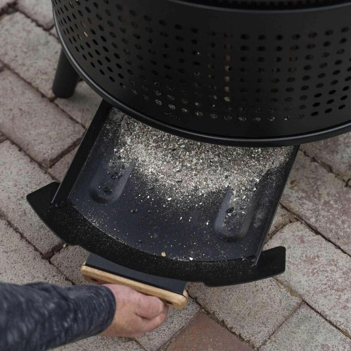 Hand pulling out a removable ash pan from a TIKI® fire pit for easy cleaning on a stone patio.