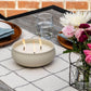 Three-wick TIKI® Brand tabletop citronella candle burning on an outdoor dining table set with flowers and place settings.