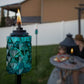 A turquoise TIKI® Brand torch burns in a backyard, adding ambiance to an outdoor seating area with a table and snacks.