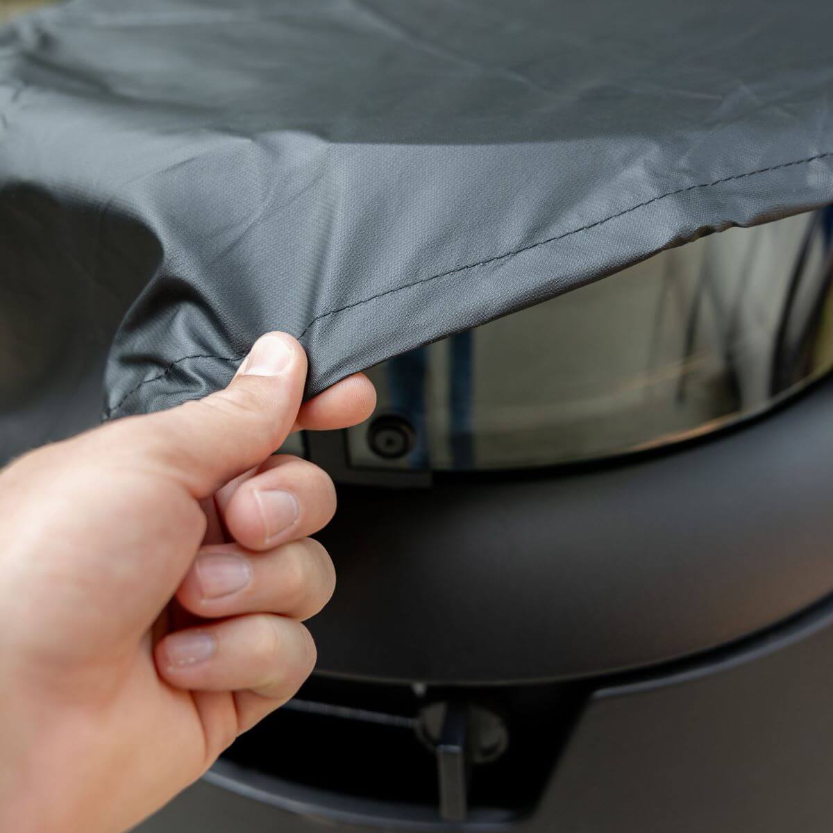 Hand placing a protective cover over a TIKI fire pit; close-up of durable, weather-resistant fabric.