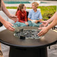 Outdoor fire pit table with glass wind guard, surrounded by people relaxing on patio furniture near a pool.