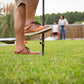 Person securing a TIKI torch stake into grass during a backyard gathering; cornhole game visible in the background.
