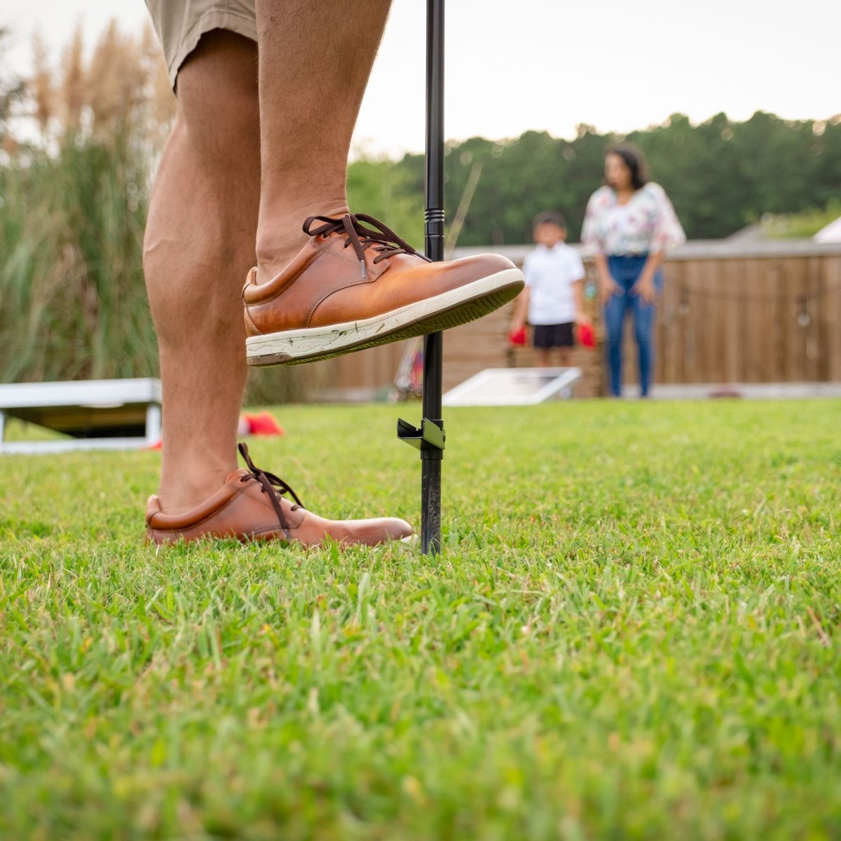 Person securing a TIKI torch stake into grass during a backyard gathering; cornhole game visible in the background.