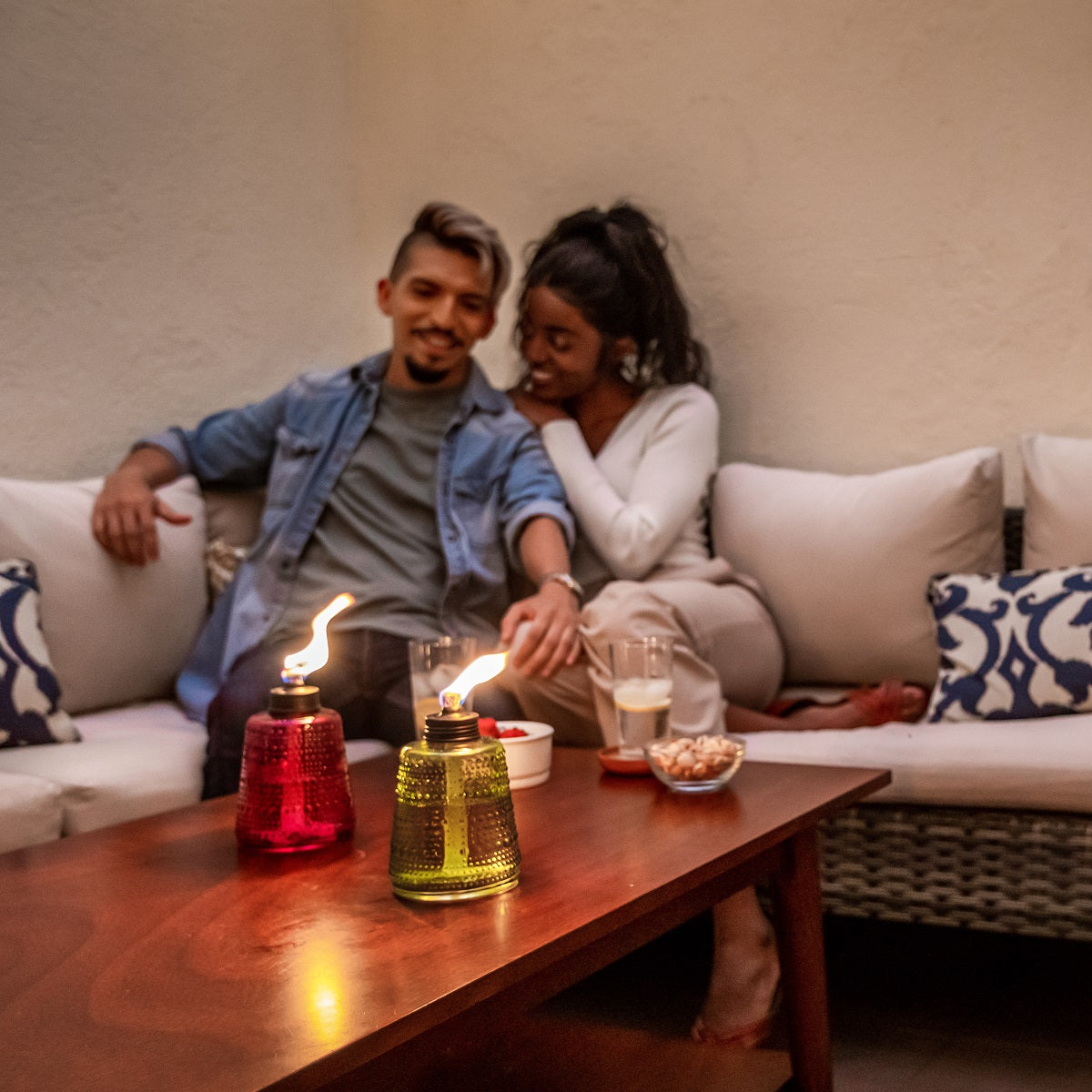 Two TIKI® Brand table torches with colorful glass bases create a cozy ambiance on a patio coffee table.