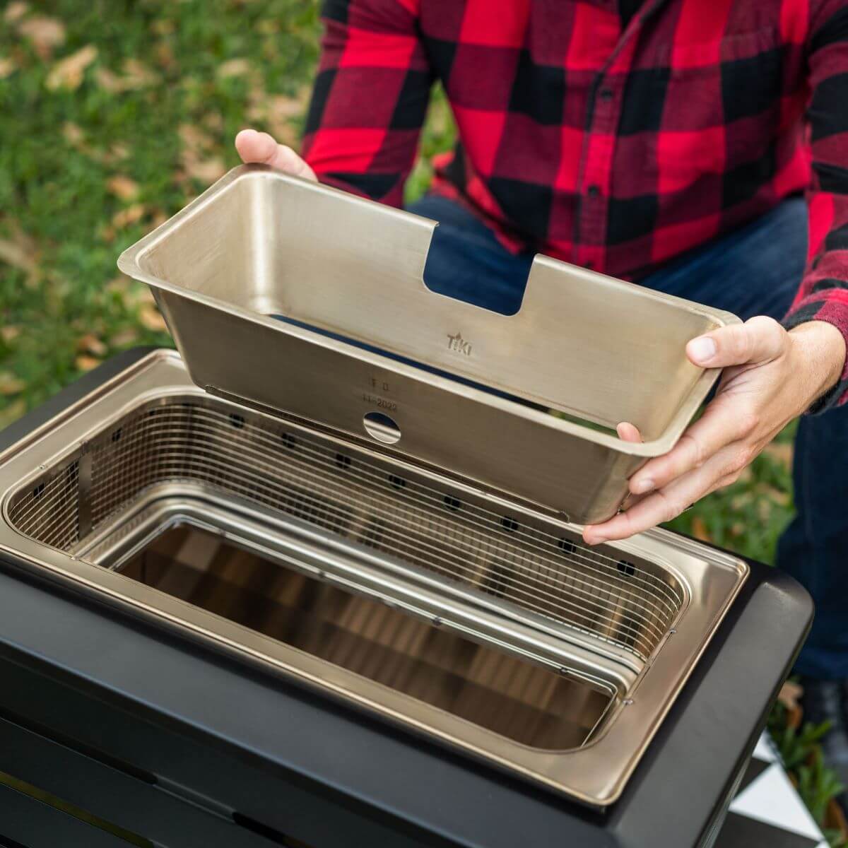 Silver TIKI drip tray for outdoor grill, held by person in red plaid shirt.