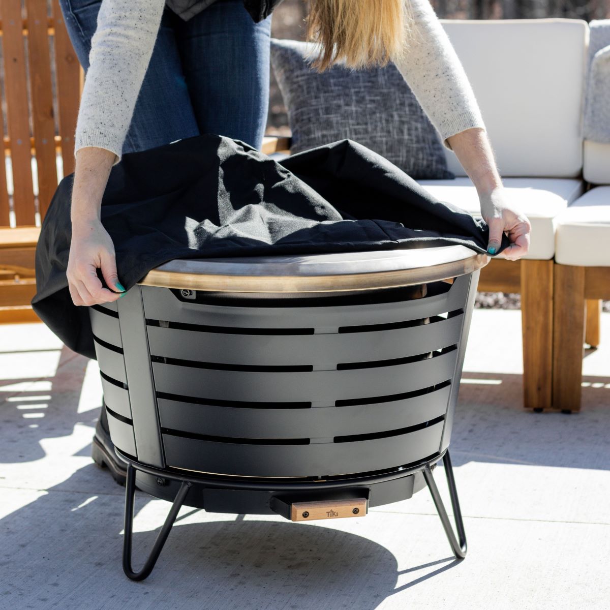 Person placing a protective cover on a TIKI fire pit on a patio, with outdoor seating in the background.