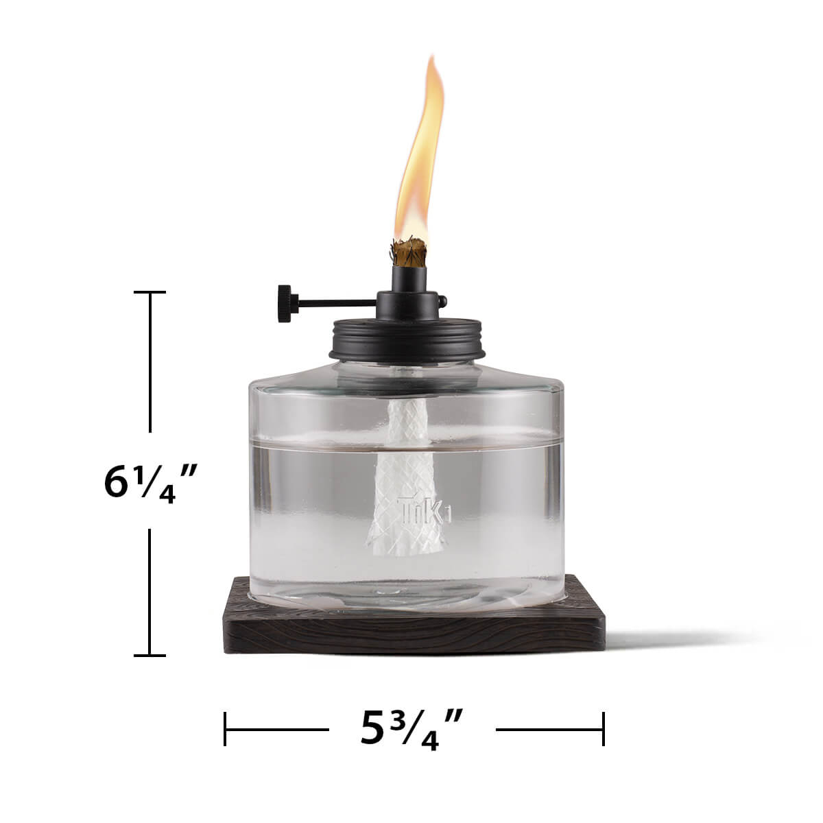 Clear glass TIKI® tabletop torch with flame, 6.25" tall and 5.75" wide, shown on a dark wood base.