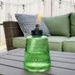 Green TIKI glass tabletop torch with flame, placed on outdoor patio table near wicker sofa and patterned pillow.