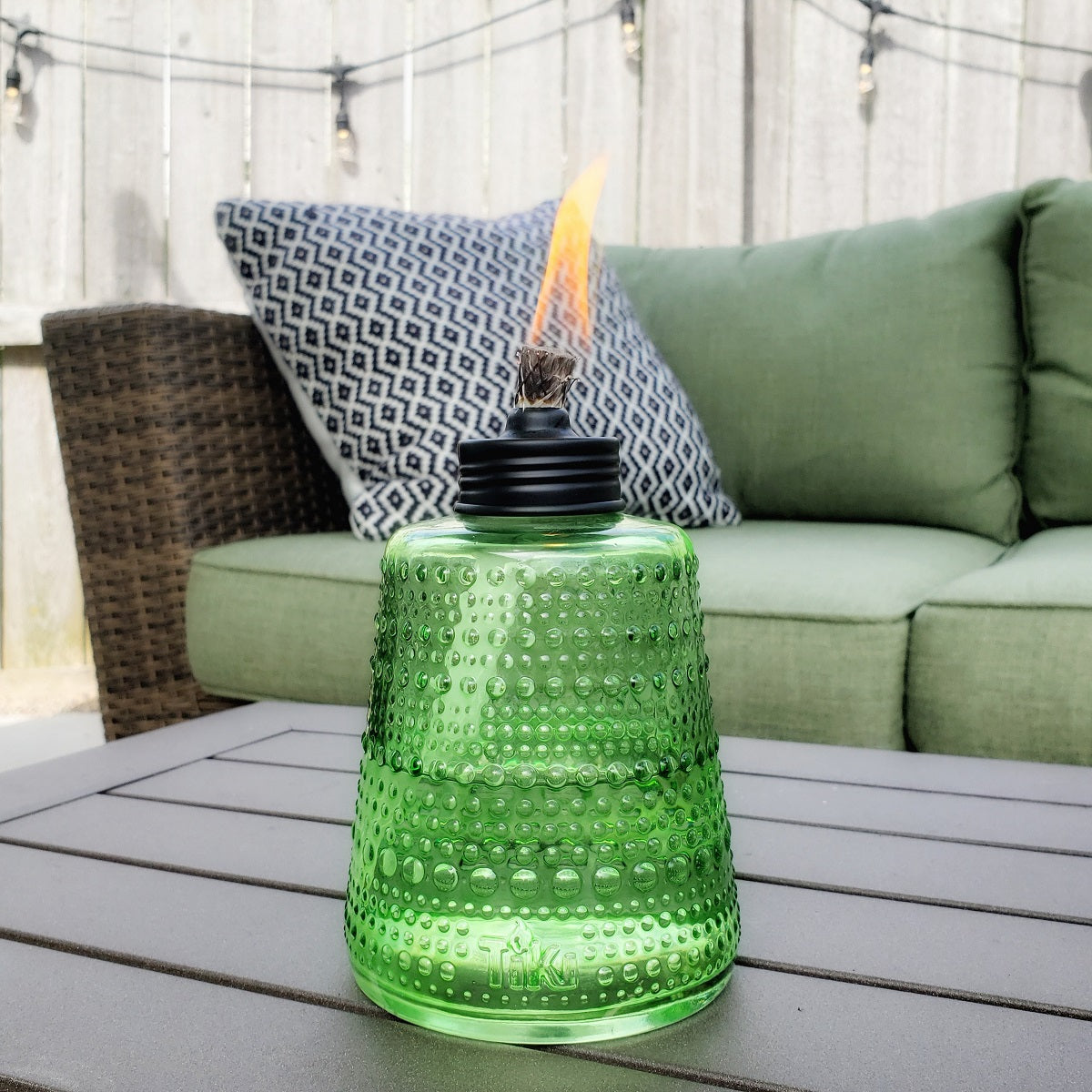 Green TIKI glass tabletop torch with flame, placed on outdoor patio table near wicker sofa and patterned pillow.