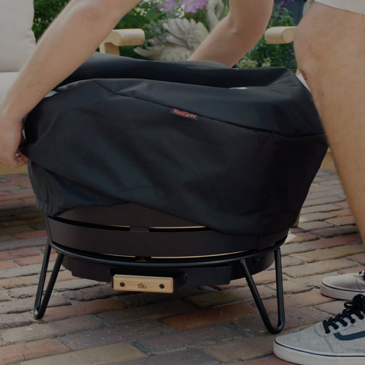Person placing a protective cover over a TIKI fire pit on a brick patio; accessible outdoor product.