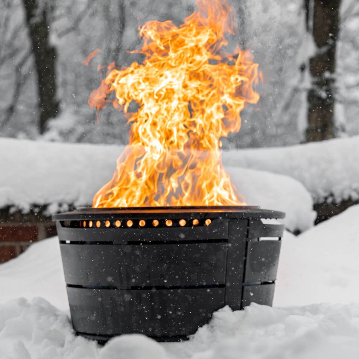 TIKI fire pit burning brightly outdoors in snowy winter setting; black metal design stands out against white snow.
