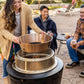 Group enjoying drinks outdoors while assembling a TIKI fire pit on a patio, with snacks on a nearby table.