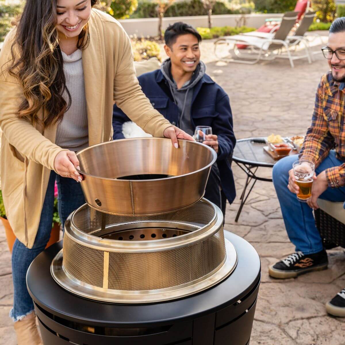 Gold mesh grill basket on black outdoor grill, surrounded by people enjoying food and drinks.