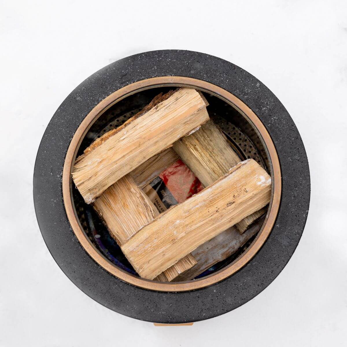 Top view of a TIKI fire pit with stacked firewood, ready for lighting. Black circular rim on a white background.
