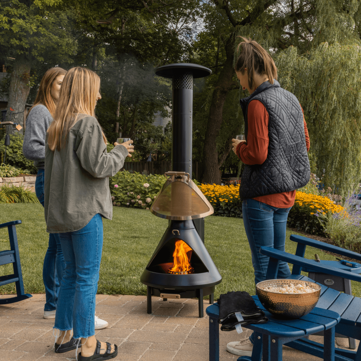 Three people gather around a TIKI Brand outdoor fire pit in a backyard, enjoying drinks and snacks on a patio.